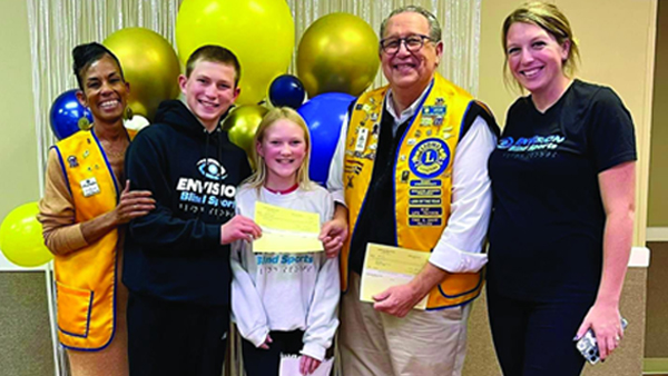 Lions club members and two young participants pose together in front of yellow and blue balloons during a club event.
