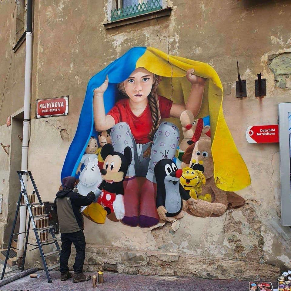 A guy painting a wall with an image of a child hiding under a Ukrainian flag